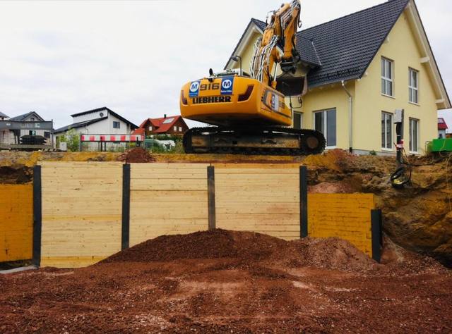 Kettenbagger auf einem Holzverbau an einer Baugrube neben einem Wohnhaus